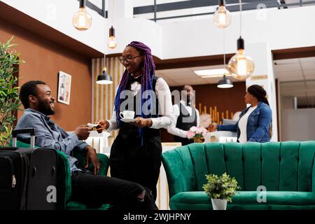 Overjoyed satisfied African American man tourist giving cash money to waitress girl while sitting in hotel lobby with luggage. Traveler buying coffee while waiting for guest check-in procedure Stock Photo