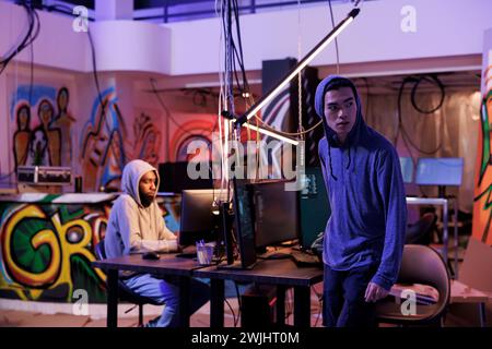 Suspicious hacker looking away after breaking into server and doing illegal activity. Young asian criminals in hood leaving abandoned warehouse after breaching database and stealing data Stock Photo