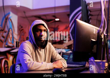 African american hacker doing illegal activities on computer and working late portrait. Criminal in hood breaking website with ddos attack and looking at camera in abandoned warehouse Stock Photo