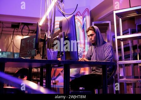 Asian hacker doing phishing attack while using illegal ransomware. Young internet criminal in hood installing malware for password cracking on victim computer in dark abandoned warehouse Stock Photo