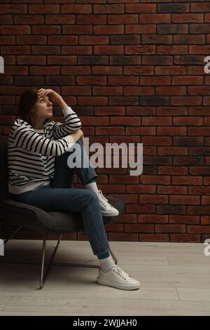 Sad young woman sitting on chair near brick wall indoors Stock Photo