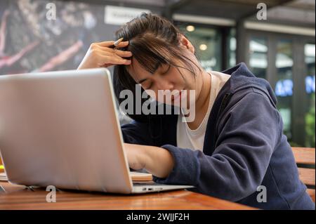 A stressed young Asian woman is worried about her project's deadline and feeling tired while working on her laptop computer at a cafe. Stock Photo