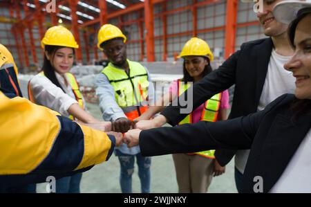 Group of construction workers clad in hard hats and reflective vests, are locked in a cordial fist bump, signaling joint progress. Machinery and steel Stock Photo