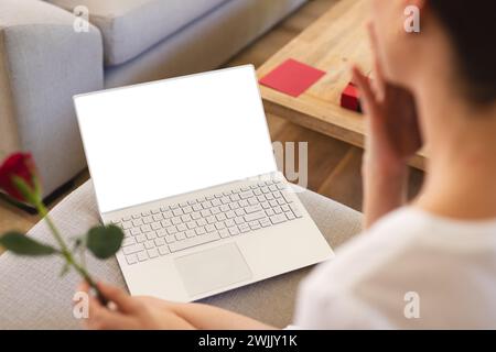 A young woman is seen from behind holding a rose while looking at a laptop screen, with copy space Stock Photo