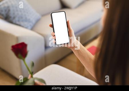A young Caucasian woman is holding a smartphone with a blank screen, with copy space, and a red rose Stock Photo