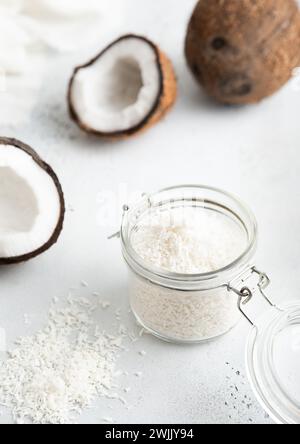 Glass jar with shredded sweet coconut flakes and ripe coconuts on light table. Stock Photo