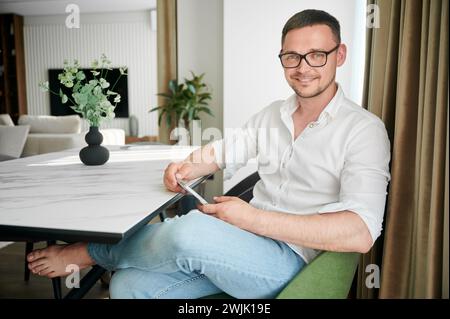 Portrait of man engaging with smartphone sitting at white marble countertop on kitchen, wearing glasses. Smiling man relaxing in his apartment. Stylish man looking straight on camera. Stock Photo