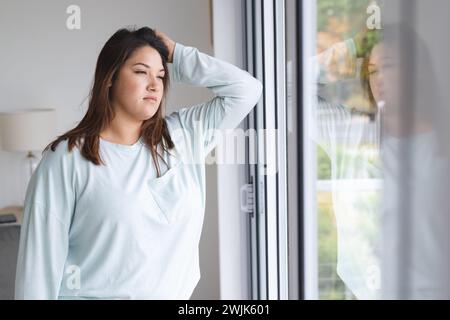 Young plus size biracial woman looks contemplative by a window at home Stock Photo