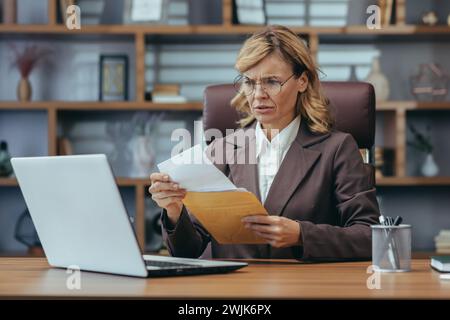 Focused senior business woman working from a home office, examining documents with a concerned expression in a modern interior. Stock Photo