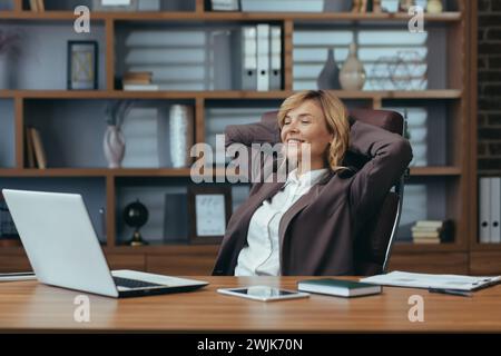 Satisfied mature businesswoman with a pleasant smile taking a break in the office, showcasing success and comfort at work. Stock Photo