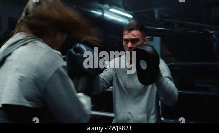 Close up of athletic woman hitting punch mitts on boxing ring. Female boxer in boxing gloves exercises with male coach and prepares to fighting competition in gym. Physical activity and workout. Stock Photo