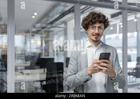A young businessman with curly hair checks his smartphone in a well-lit, contemporary office environment, exuding professionalism and focus. Stock Photo