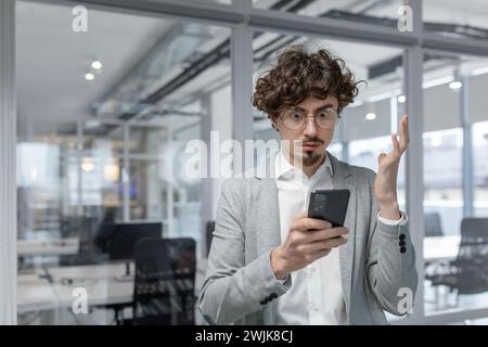 Curly-haired young adult businessman looks confused while using a mobile phone in a modern office environment. Stock Photo