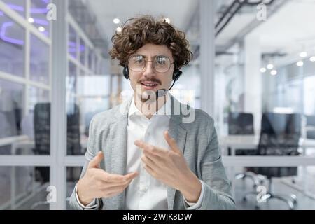 Curly-haired mature businessman with headset actively gestures while communicating in a brightly lit office environment. Stock Photo