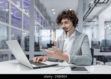 Confident young adult businessman with curly hair using laptop and headset in an office setting, engaging in corporate work. Stock Photo