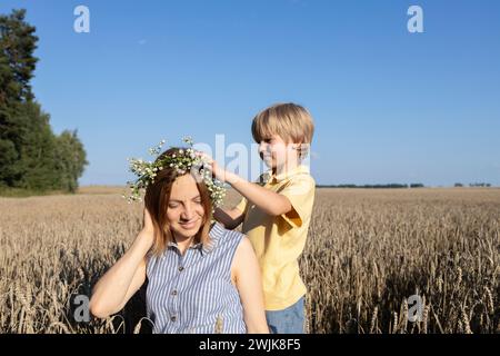 happy family, mother and son in a wheat field. The concept of parental love, motherhood. the boy puts a wreath of daisies on his mothers head. enjoy n Stock Photo