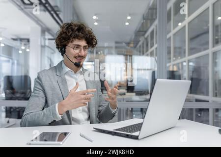 Curly-haired businessman converses on headset while working on laptop in a modern office setting, showing professionalism and expertise. Stock Photo