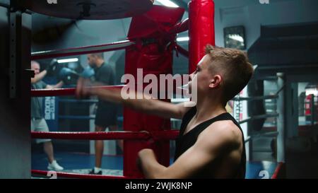 Focused young fighter hits punching bag during workout in dark boxing gym. Athletic teenage boy exercises before fighting competition or tournament. Boxer prepares to fight and trains. Back view. Stock Photo