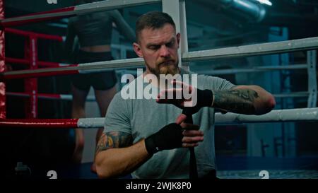 Adult man sits near boxing ring and wraps his hands with bandage for martial arts. Athlete prepares to fight, competition or training in box gym. Female boxer performs shadow boxing at background. Stock Photo