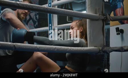 Tired boxer sits at ring corner after long fight. Male trainer comes to woman and helps her to take off boxing gloves and bandages. Athlete prepares to fight, competition or training in boxing gym. Stock Photo