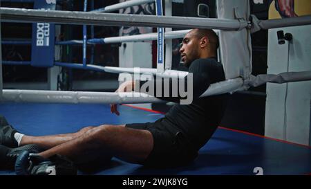 Tired and exhausted African American fighter sits in boxing ring corner. Male boxer takes off his boxing gloves and takes break during training. Athlete prepares to fight or competition in boxing gym. Stock Photo