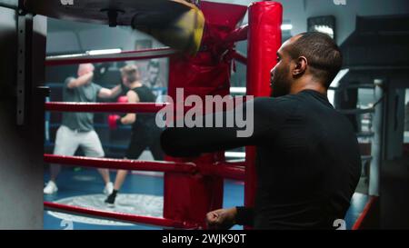 Focused African American fighter hits punching bag during workout in dark boxing gym. Athletic man exercises before fighting competition or tournament. Boxer prepares to fight and trains. Back view. Stock Photo