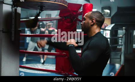 Focused African American fighter hits punching bag during workout in dark boxing gym. Athletic man exercises before fighting competition or tournament. Boxer prepares to fight and trains. Back view. Stock Photo