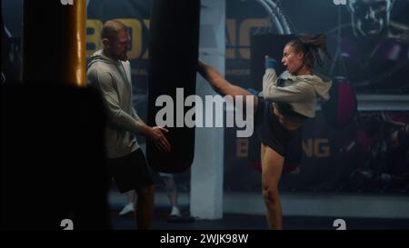 Athletic woman in boxing bandages hits punching bag while training in dark gym. Female kickboxer practices and prepares for competition with male coach. Physical activity and intensive workout. Stock Photo