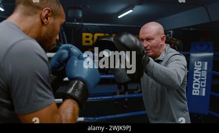 African American professional boxer in boxing gloves exercises with trainer in gym before tournament. Male athlete hits punching mitts training on boxing ring. Physical activity and intensive workout. Stock Photo