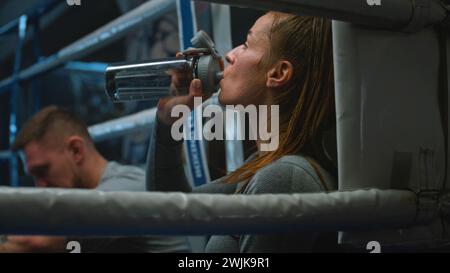 Tired woman sits at ring corner, takes off boxing gloves and starts drink water. Male trainer sits down near boxer after hard train. Female athlete prepares to fight or competition in boxing gym. Stock Photo