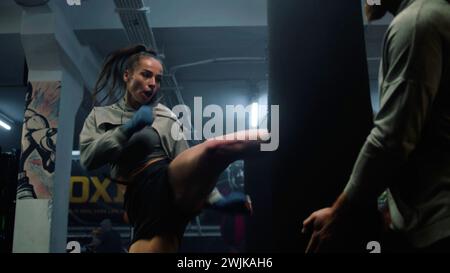 Female fighter in boxing bandages exercises with male trainer in dark boxing gym before fighting match. Athletic woman hits punching bag and practices fighting technique. Physical activity and workout Stock Photo