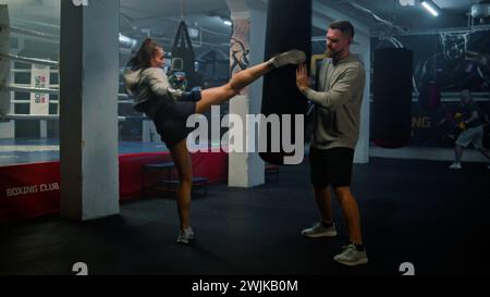 Female kickboxer in boxing bandages hits punching bag while training in dark boxing gym. Athletic woman practices and prepares for match with male coach. Physical activity and intensive workout. Stock Photo