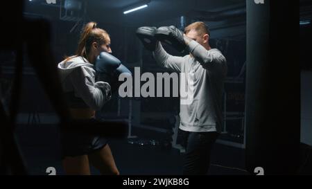 Athletic fighter in boxing gloves trains and exercises for fighting championship. Woman hits punching mitts and practices in fighting techniques with adult trainer. Physical activity and workout. Stock Photo