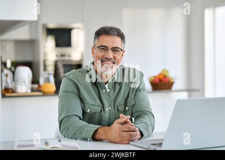 Happy middle aged man wearing glasses sitting with laptop at home table. Stock Photo