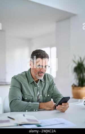 Happy middle aged man using cell phone sitting at home kitchen table. Stock Photo
