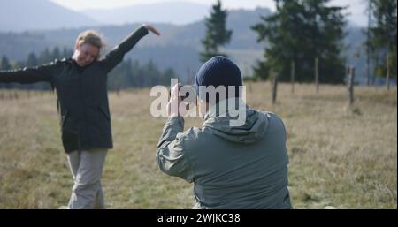 Caucasian man takes pictures of his wife on hilltop against scenic nature view using professional camera. Couple of travelers, tourist family during hiking trip or trek in mountains. Active leisure. Stock Photo