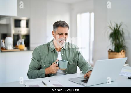 Happy older middle aged man drinking coffee using laptop at home table. Stock Photo