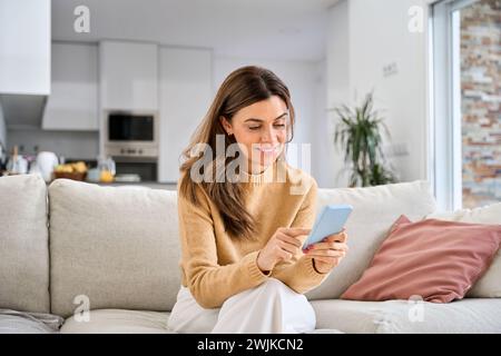 Happy mature older woman using smartphone sitting on couch at home. Stock Photo