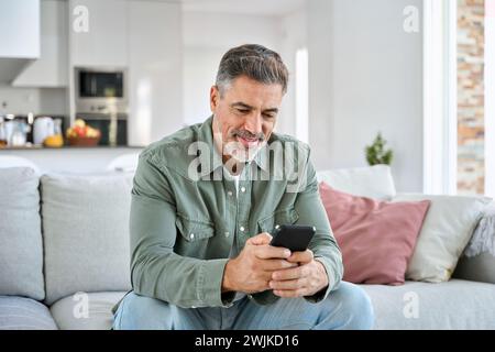Middle aged old man using smartphone relaxing on couch at home. Stock Photo