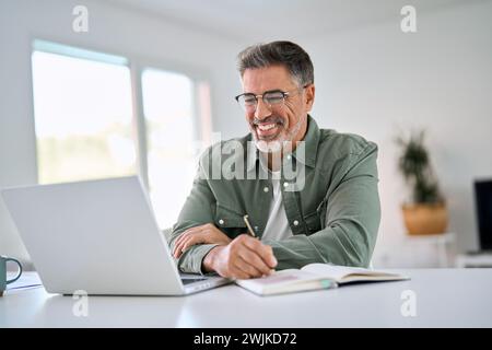Happy middle aged man using laptop computer writing notes sitting at home table. Stock Photo