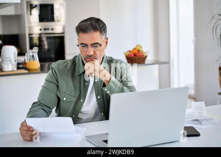 Worried mature man looking at bill working at home kitchen using laptop. Stock Photo