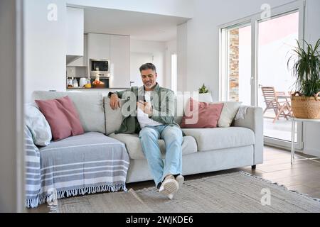 Smiling relaxed middle aged man using mobile phone sitting at home on couch. Stock Photo