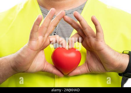 Close-up of builder man in fluorescent vest holding red heart symbol making love gesture using fingers isolated on white background Stock Photo