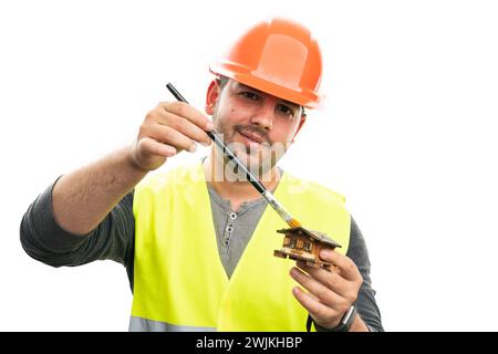Constructor man wearing orange helmet and fluorescent vest painting miniature wooden house using brush smiling isolated on white background Stock Photo