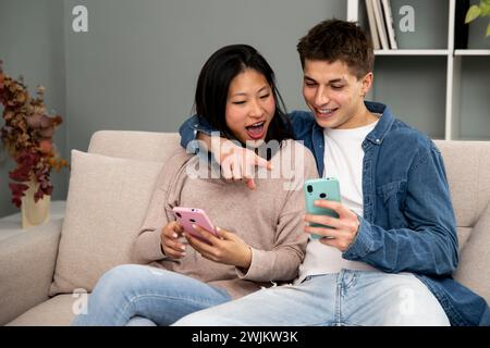 Affectionate young couple with mobile phones relaxing on sofa. Happy diverse woman and man hugging using smartphone together sitting on cozy couch at Stock Photo