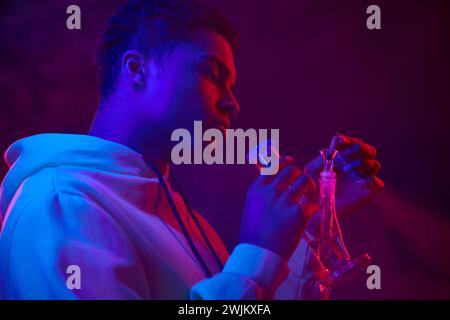 young african american man in hoodie lighting glass bong on dark blue background with red light Stock Photo