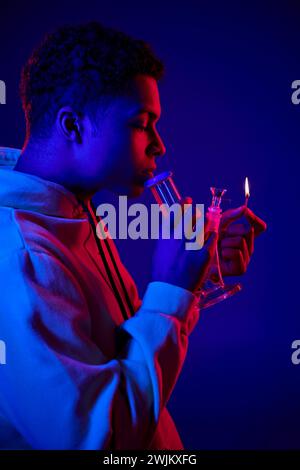 side view, african american man in hoodie lighting glass bong on dark blue background with red light Stock Photo