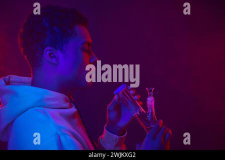 young african american man in hoodie looking at glass bong on dark purple background with lighting Stock Photo