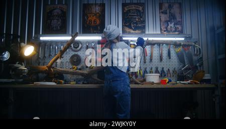 Creative female carpenter comes in workshop, wears protective glasses and starts to grind tree branch using grinder machine. Young woman artisan woodworking, makes wooden products. Handmade concept. Stock Photo