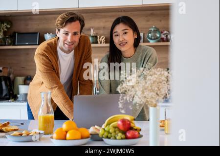 Adult couple looking at the camera while using laptop at kitchen counter Stock Photo